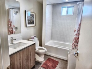 Ein Badezimmer mit weißem Waschbecken, WC und Badewanne. An der Wand hängt ein gerahmtes Bild, und der Duschvorhang ist mit floralen Mustern versehen.