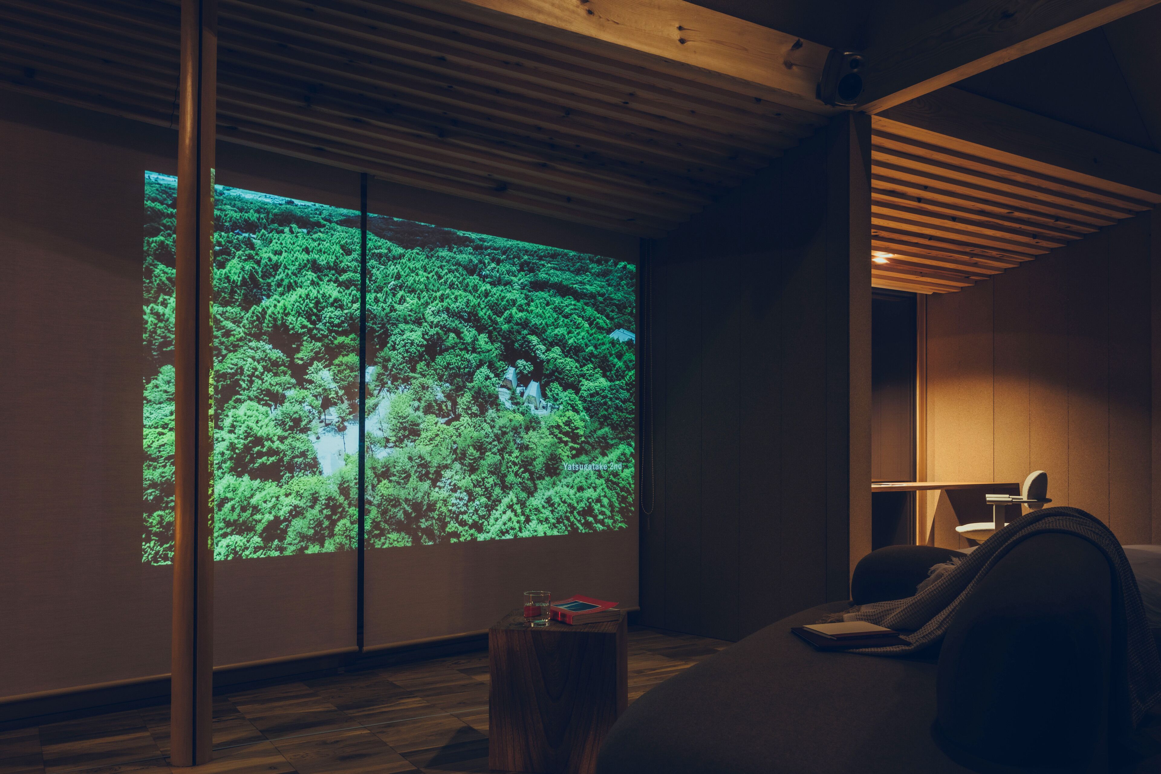 A room with a large projection screen displaying a forest scene, a wooden ceiling, a small wooden table with a glass and a book, and a bed with a blanket.