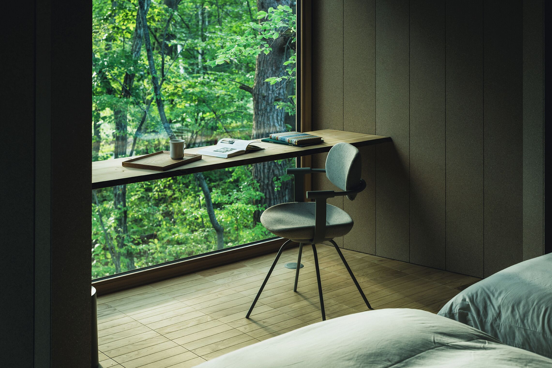 A room with a large window overlooking a forest, a desk with a chair, and a bed.