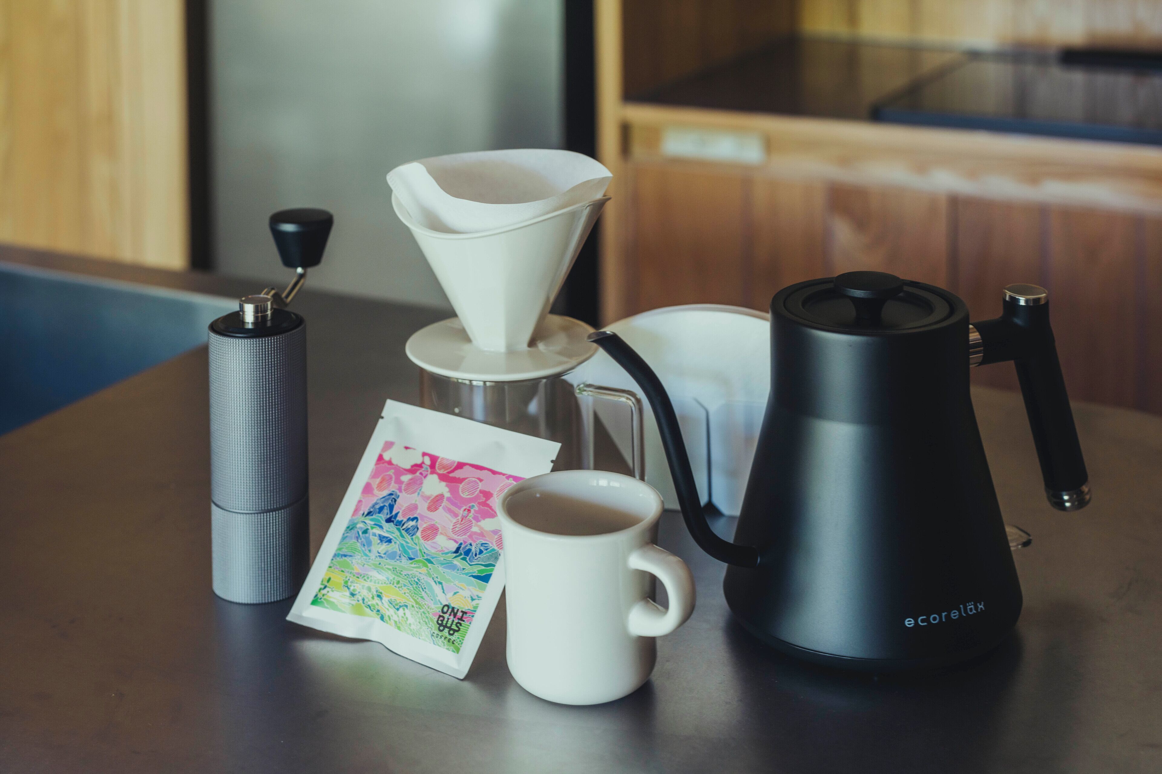A coffee maker, a white mug, a silver container, and a colorful card on a wooden table.