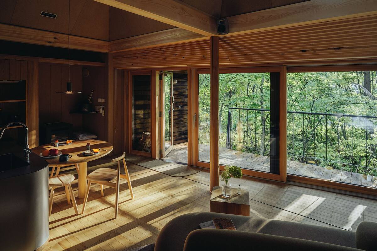 L'intérieur d'un chalet moderne est doté d'un plancher en bois, d'un coin repas avec des chaises, d'une cuisine avec un poêle et de grandes fenêtres offrant une vue sur la forêt.