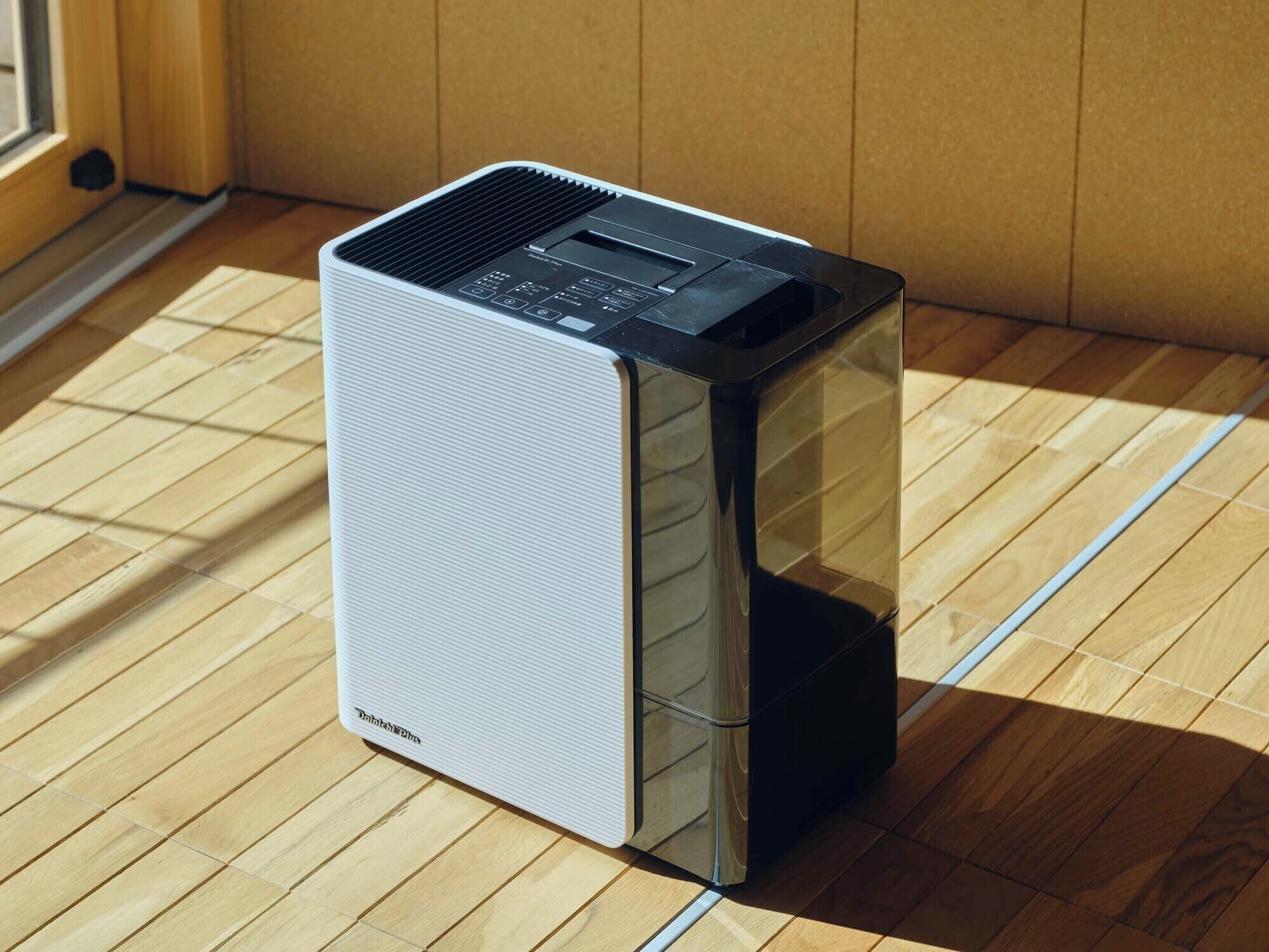A compact, modern air purifier with a transparent front panel, placed on a wooden floor near a window.
