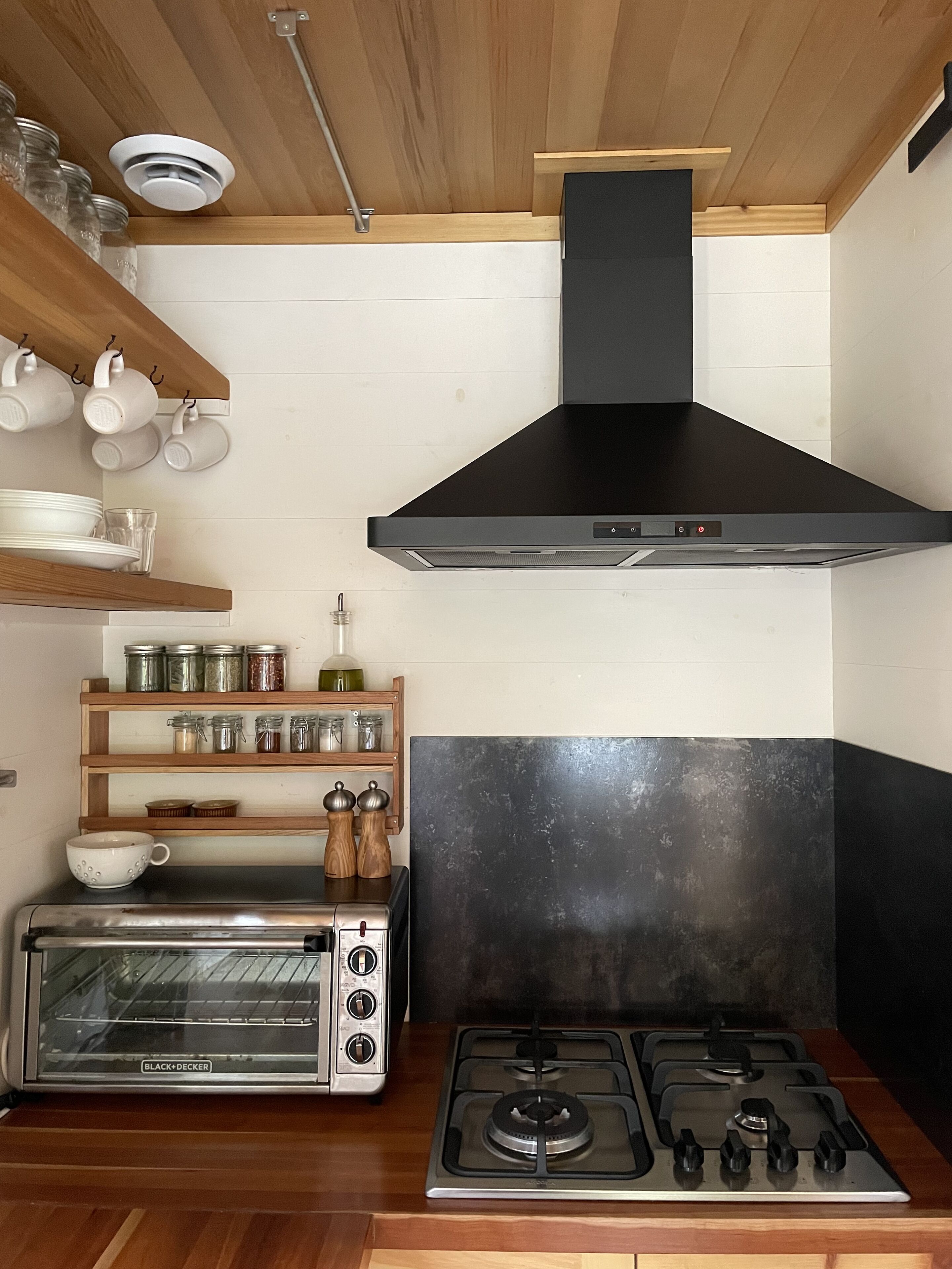 A kitchen with a black range hood, a built-in oven, and a gas stove. There are shelves with jars and a toaster oven.