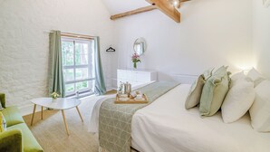 Una habitación con techo inclinado, una cama con almohadas, una mesita de noche con una bandeja, una pequeña mesa redonda, un sofá y una ventana con cortinas.