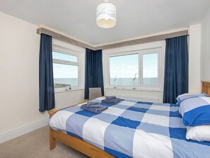 Une chambre à coucher avec une couvre-lit à carreaux, deux fenêtres avec des rideaux, une vue sur la mer et un luminaire suspendu.