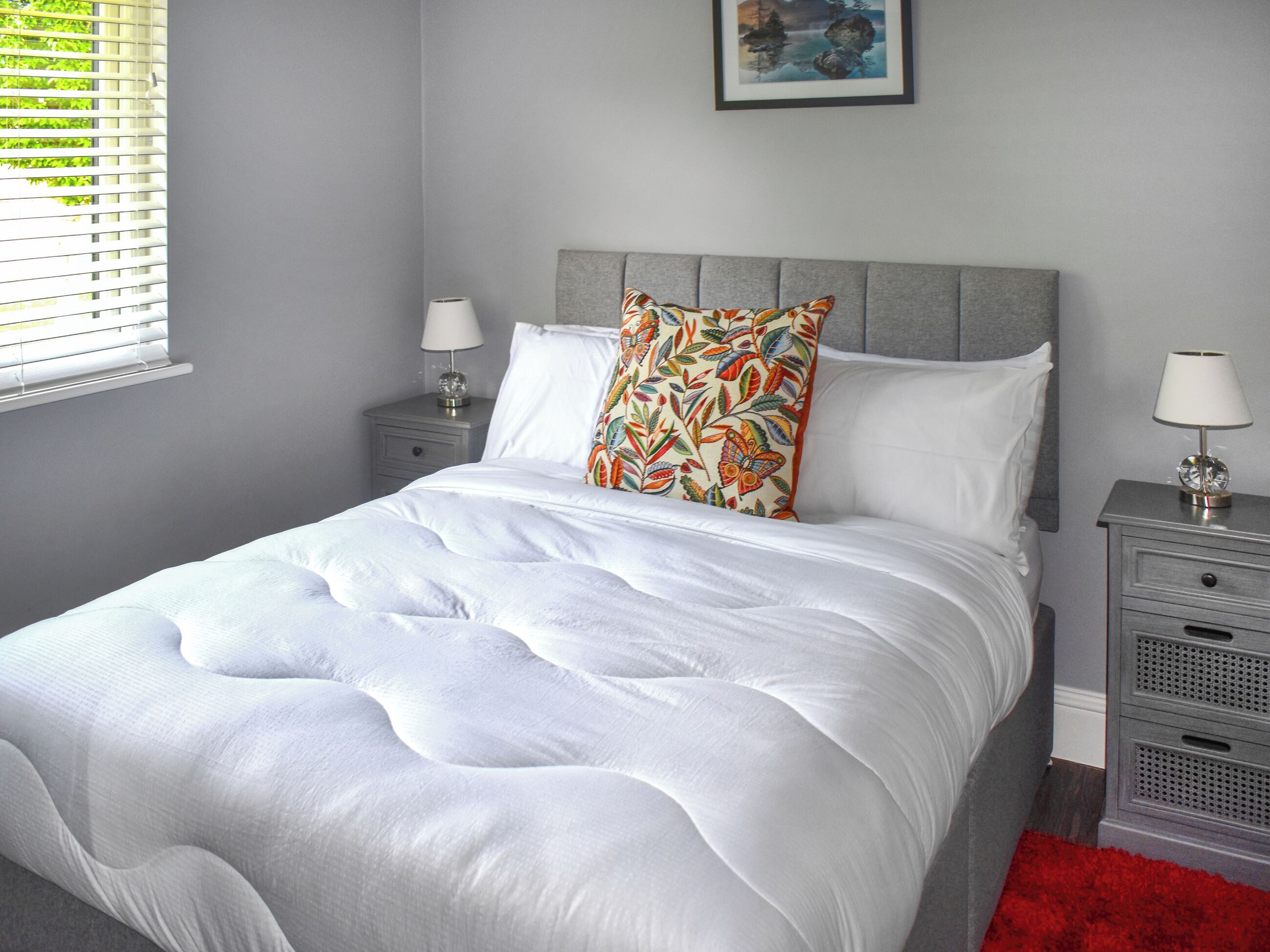 Una cama bien hecha con ropa blanca y una almohada colorida, dos mesitas de noche con lámparas, un cuadro en la pared y una ventana con persianas.