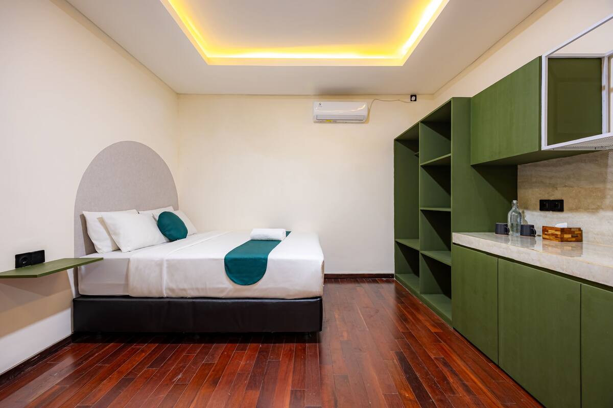 Una habitación de hotel moderna con cama, armarios verdes, una pequeña cocina y una lámpara de techo.