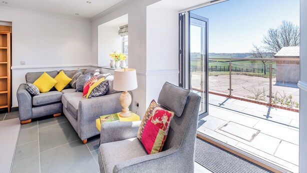 A modern living room with grey sofas, yellow and red cushions, a white lamp, and a large glass door leading to an outdoor patio. - The Stables - UK49089 (Crediton)
