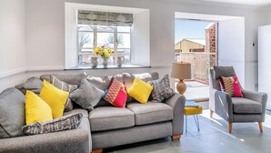 A modern living room with a grey sectional sofa, yellow and patterned cushions, a side table with a lamp, and a view of the outside through large windows. - The Stables - UK49089 (Crediton)