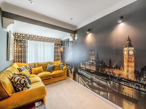 Una habitación con un gran mural de un paisaje urbano, un sofá amarillo y una ventana con cortinas.
