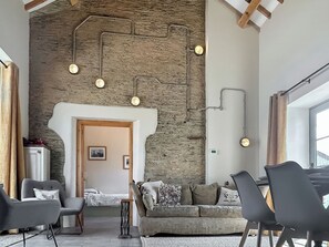 A modern living room with a stone wall, a wooden door, and a sofa. There are two chairs and a small table. - The Tractor Barn - UK49678 (Bratton Fleming)