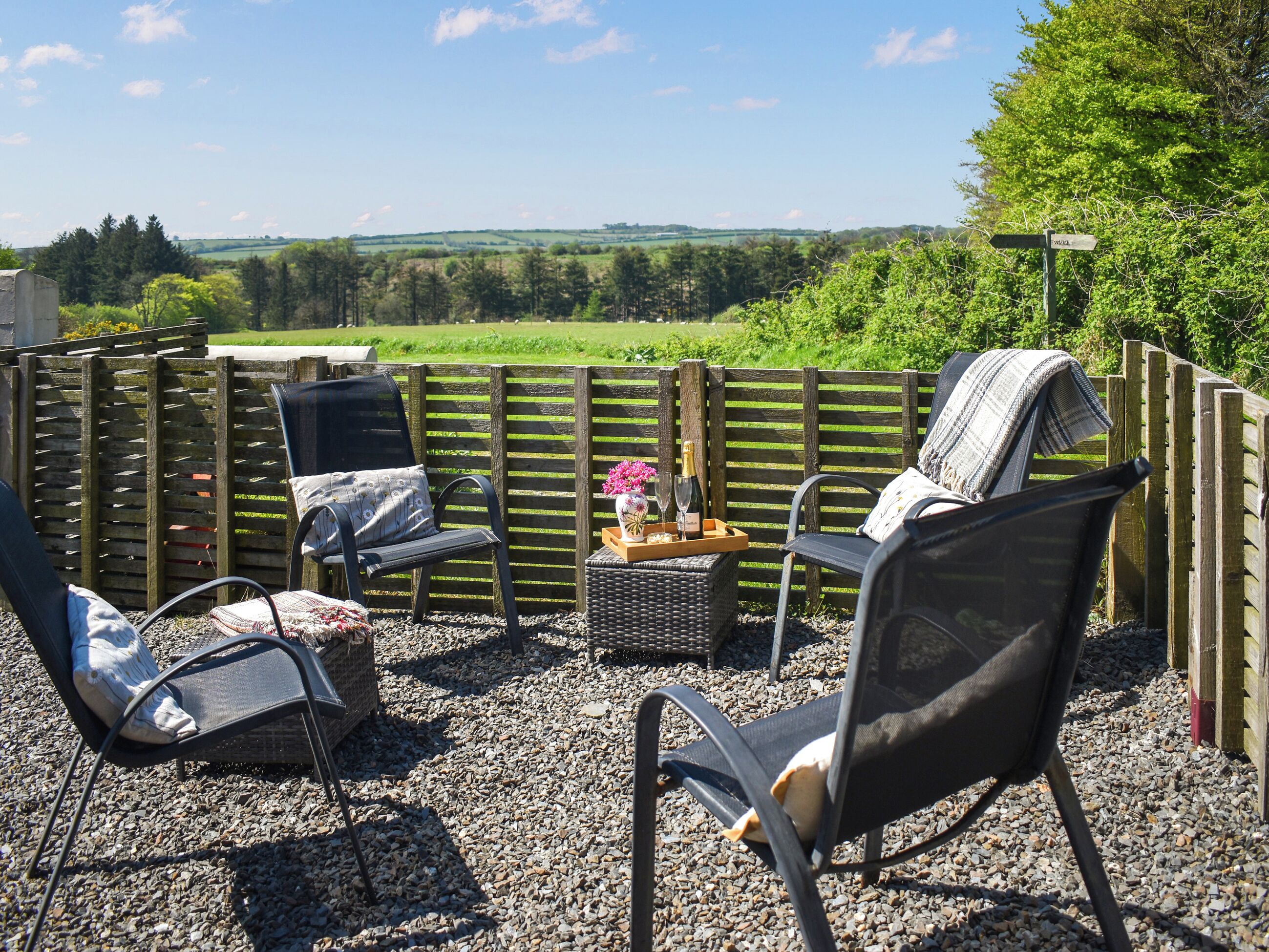 Un espace extérieur aménagé avec des chaises et un banc en osier, une clôture en bois et une vue sur un paysage verdoyant.