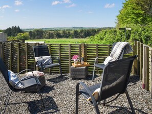 An outdoor seating area with wicker chairs and a bench, a wooden fence, and a view of a green landscape. - The Tractor Barn - UK49678 (Bratton Fleming)
