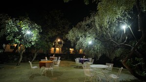 Un espace extérieur aménagé pour les repas, avec des tables et des chaises, entouré d’arbres et éclairé par des lampadaires.