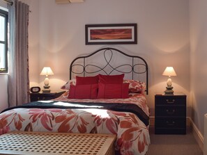 Une chambre à coucher avec un lit, deux tables de chevet avec des lampes, une commode, un tableau encadré au mur et une fenêtre avec des rideaux.