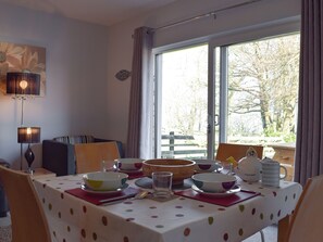 Un coin repas avec une table dressée pour quatre personnes, un lustre et une vue sur les arbres à travers de grandes fenêtres.