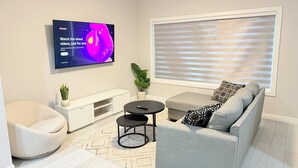Una sala de estar moderna con un sofá gris, una mesa de centro negra redonda, un televisor blanco montado en la pared y un ventanal con persianas.