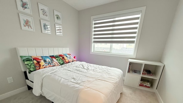 A bedroom with a bed, a window with blinds, a white headboard, and a small white shelf with books.