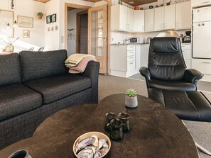 Una sala de estar con un sofá gris oscuro, un sillón de cuero negro, una mesa redonda de centro con una planta, vasos y un bol de aperitivos.