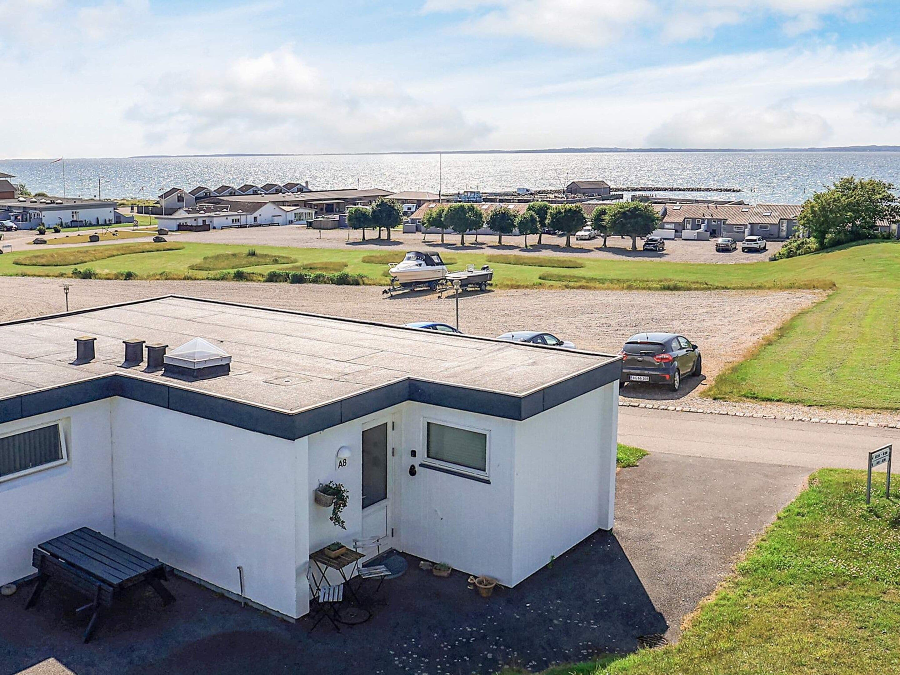 Un edificio pequeño de un piso con techo plano, un patio con mesa y sillas, y vistas a un cuerpo de agua y un puerto deportivo al fondo.