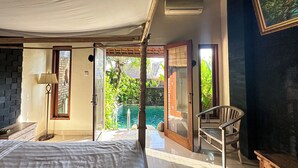 Un dormitorio con cama, mesita de noche, lámpara, una silla de madera y vistas a una piscina y zonas verdes exteriores.