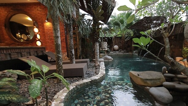 Un área de piscina interior tranquila con pared de ladrillo, un espejo circular y diversas plantas.