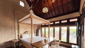 Una cama con dosel de madera y ropa de cama blanca, una pequeña mesa redonda con una silla, y un ventanal con vistas a edificios y vegetación.