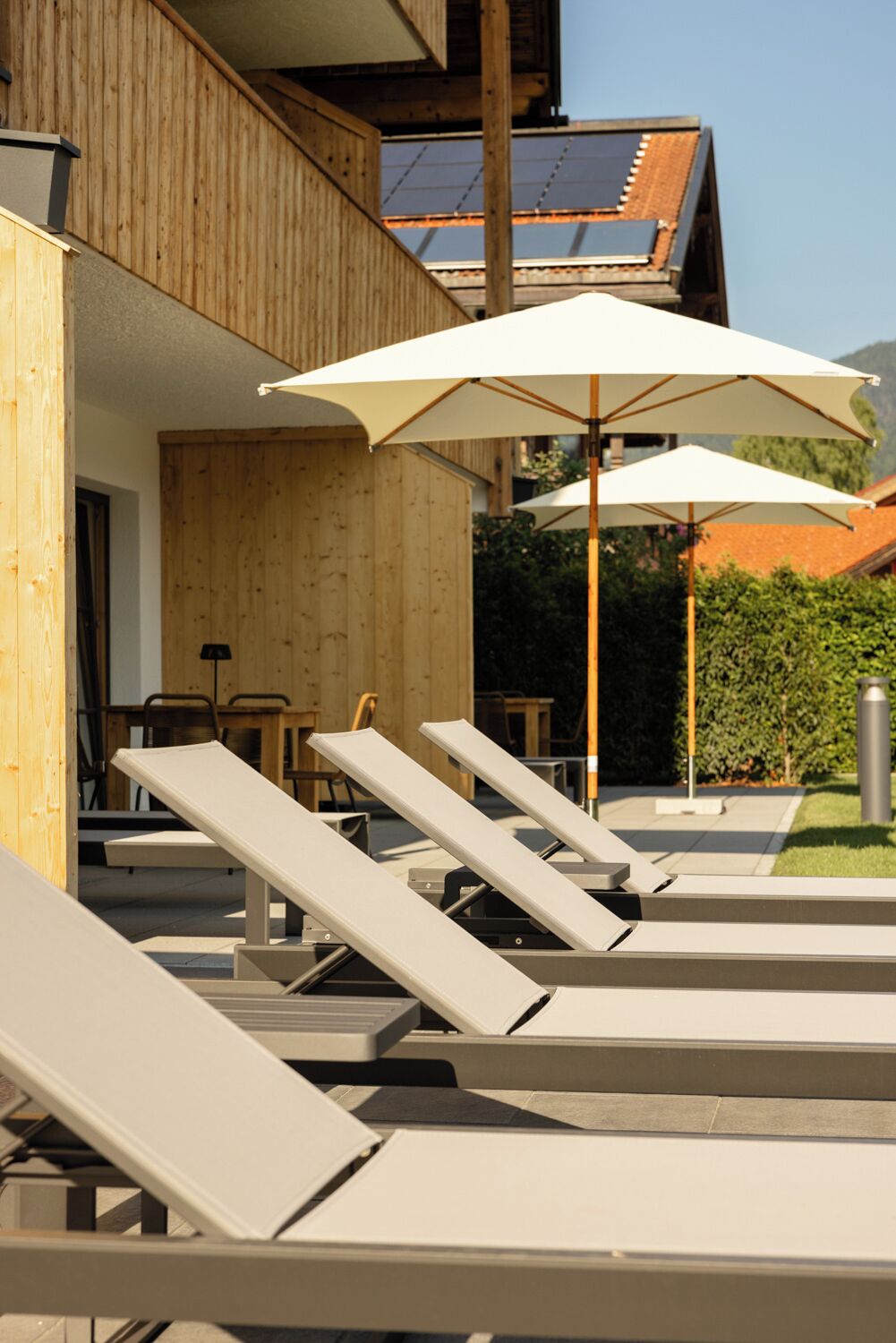 A modern outdoor lounge area with sun loungers and umbrellas, wooden walls, and a view of a building with solar panels.