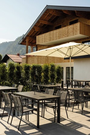 An outdoor seating area with tables and chairs, a wooden building with a balcony, and a mountain backdrop. - 3-room vacation apartment, 29 m², with modern furnishings (Reit im Winkl)
