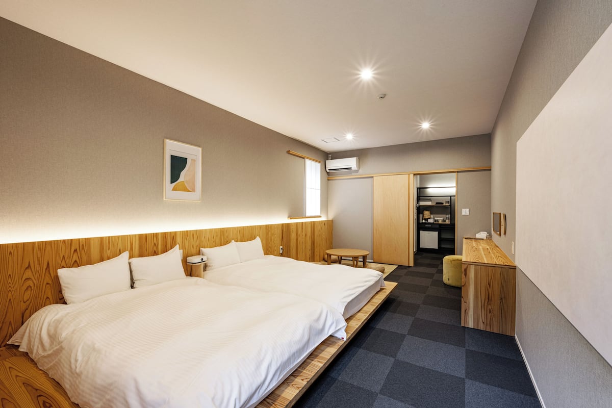 A modern hotel room with two beds, a small table, and a kitchenette in the background.