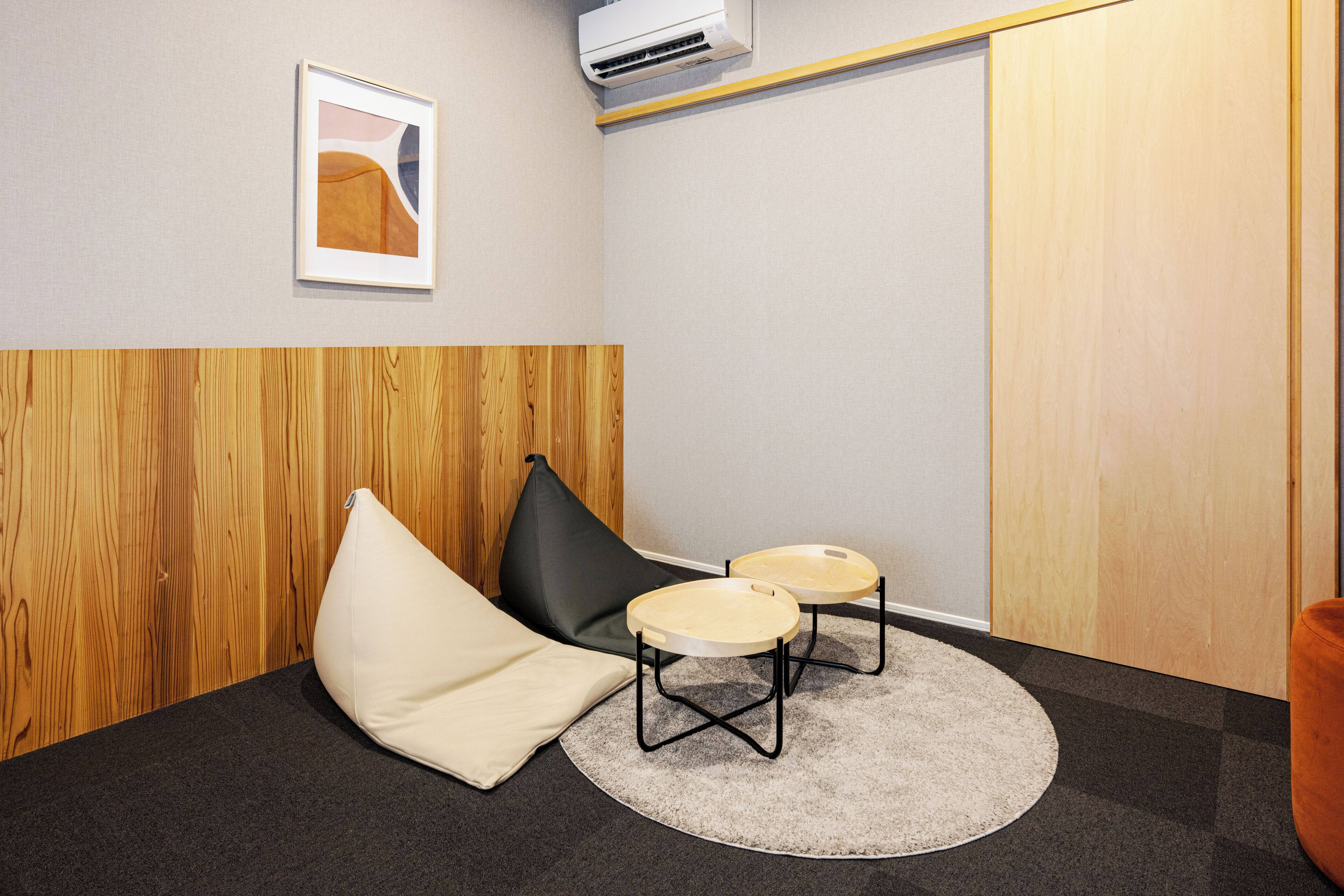 Ein Zimmer mit einer Wandpaneele aus Holz, einem gerahmten abstrakten Kunstwerk, einem schwarzen Sitzsack, zwei runden Tischen mit schwarzen Metallbeinen und einem grauen Teppich.