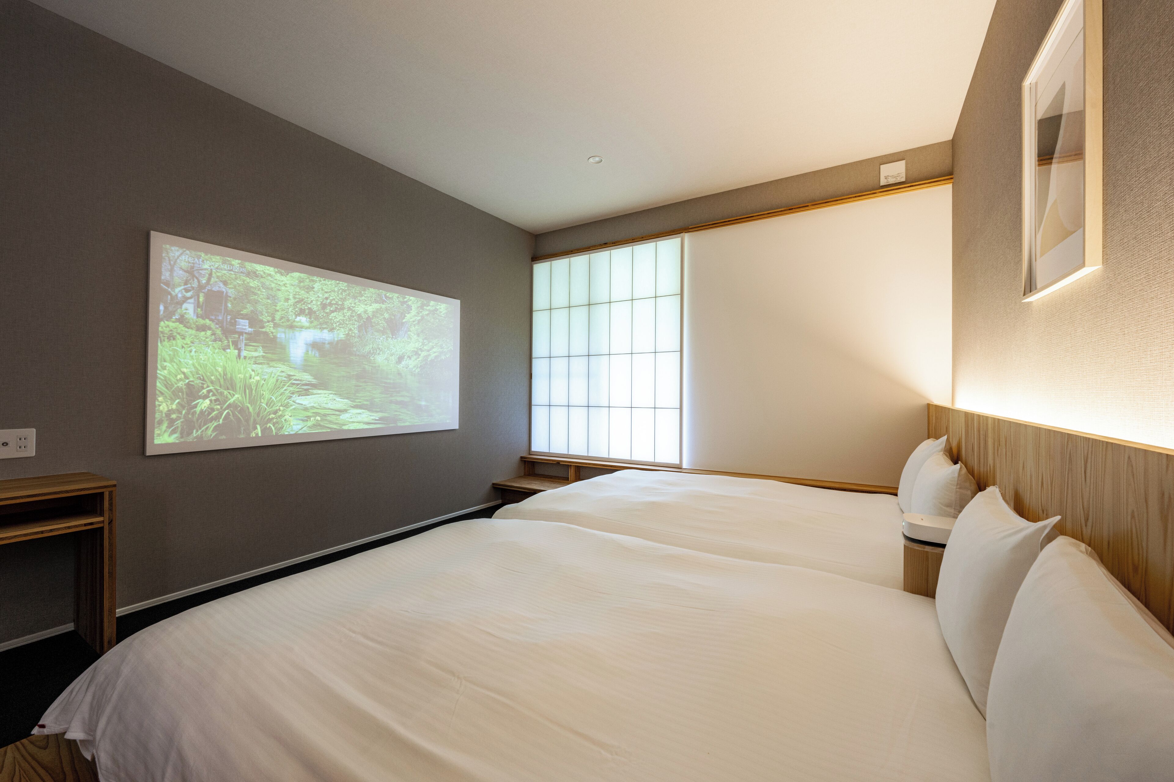 A hotel room with a bed, a projector screen displaying a nature scene, a window with blinds, and a wooden headboard.