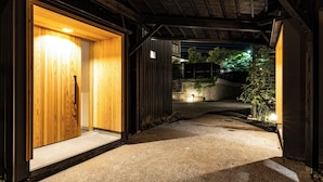 A pathway leading to a wooden door with a small window, surrounded by a concrete area with a metal fence and a small tree. - Beppu Hotel Negura (Beppu)