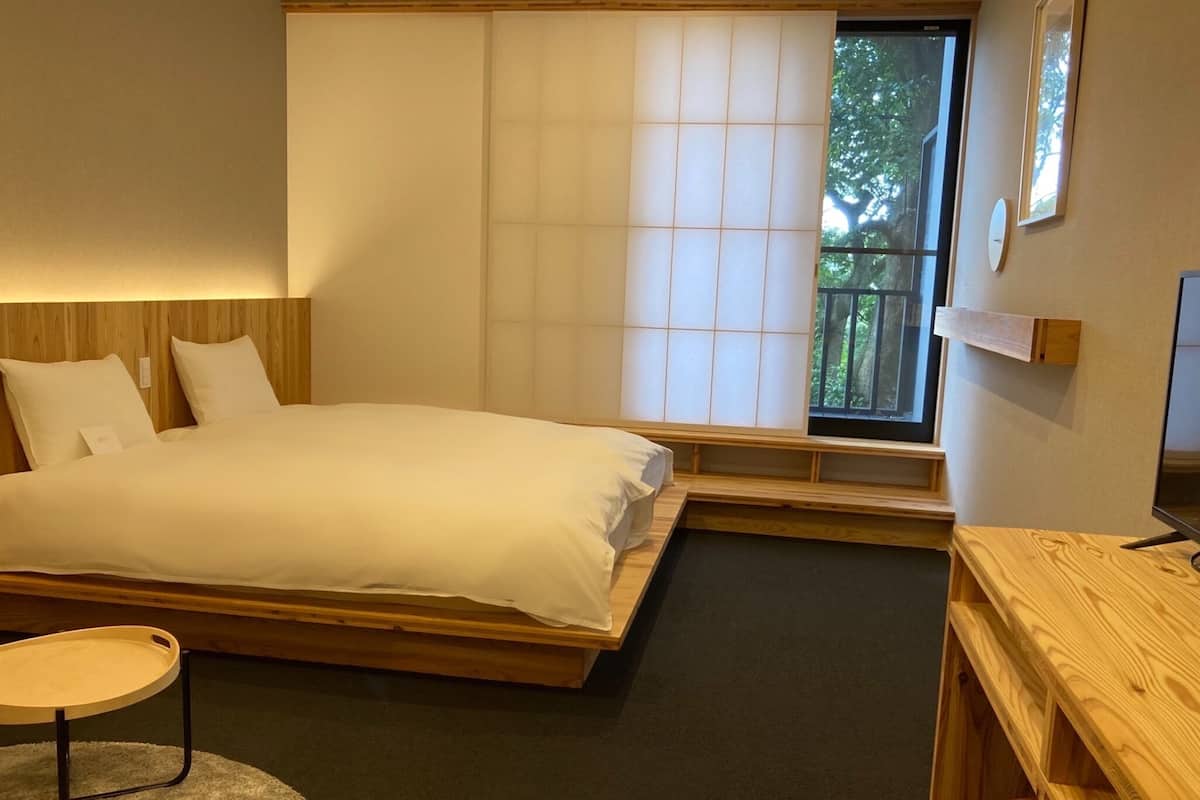 A bedroom with a bed, a wooden desk, a chair, and a window with a view of trees.