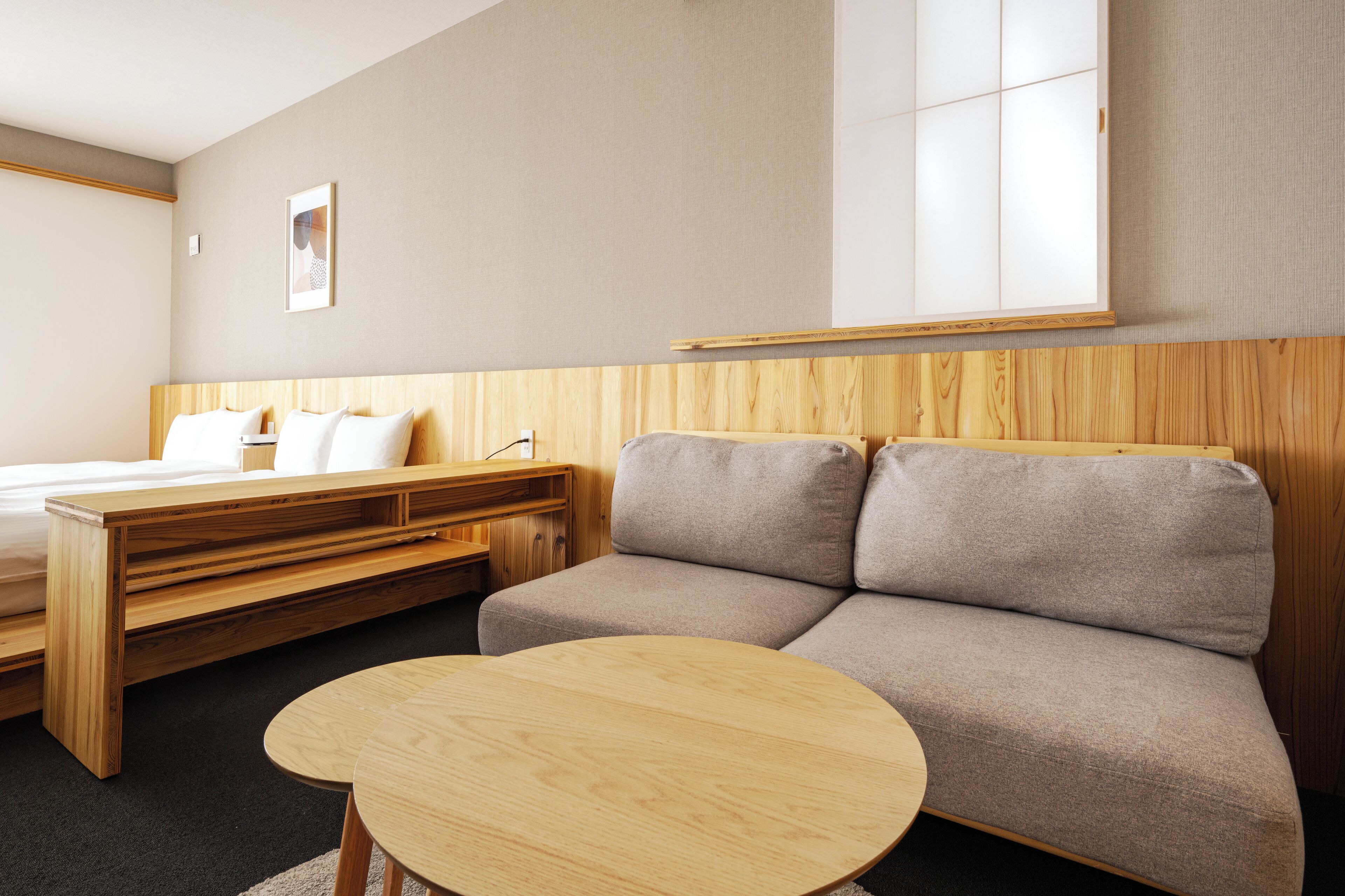 A modern hotel room with a bed, a sofa, a small wooden table, and a window with a view.