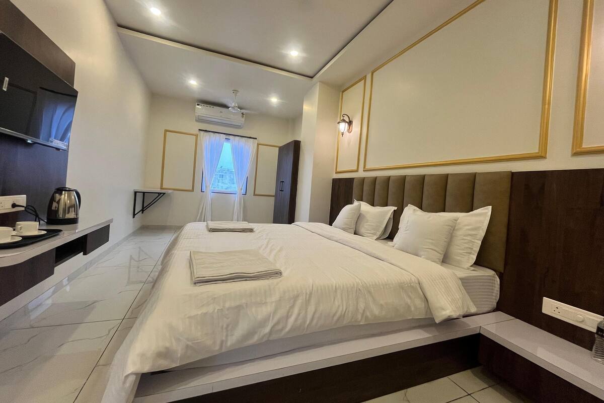 A modern hotel room with a large bed, bedside tables, a desk with a kettle, and a window with curtains.