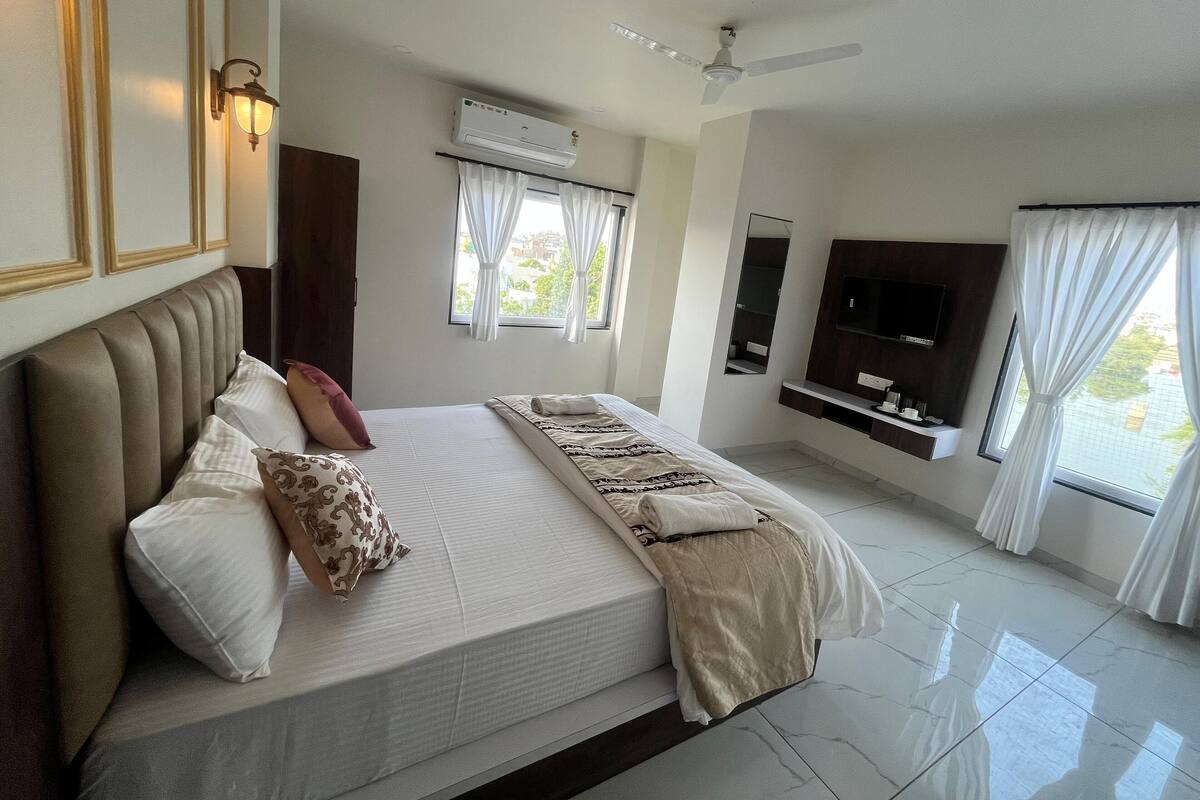 A hotel room with a large bed, a ceiling fan, an air conditioning unit, a TV mounted on the wall, and a window with curtains.