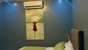 A bedroom with a bed, green and white bedding, a headboard, a wall-mounted air conditioner, and a painting of a woman with an umbrella. - Spacious 3-bedroom hotel suites with AC, WiFi in enjoyable City of Santa Rosa (City of Santa Rosa)