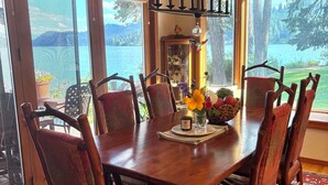 Un coin repas avec une table en bois, des chaises et un lustre. De grandes fenêtres offrent une vue sur un lac et des arbres.