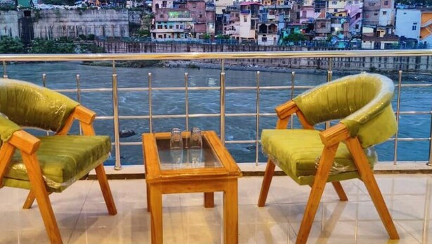 A balcony with two green chairs and a small table, overlooking a river with colorful buildings in the background. - Hotel Ganga Sagar (Bhatwari)