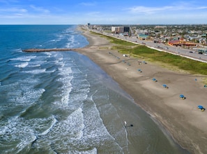 Vista aérea de una playa con una extensa franja de arena, un muelle que se adentra en el mar y un paisaje urbano al fondo.