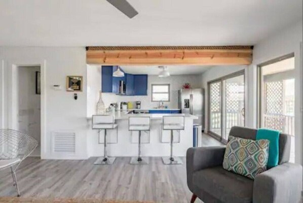A modern kitchen with white cabinets, a central island, and a bar area. There is a gray sofa with a patterned pillow in the living area.