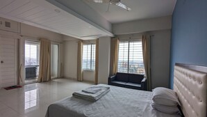 A hotel room with a bed, a chair, a ceiling fan, and large windows with curtains. - YMCA Training Center and Guest House  (Savar)
