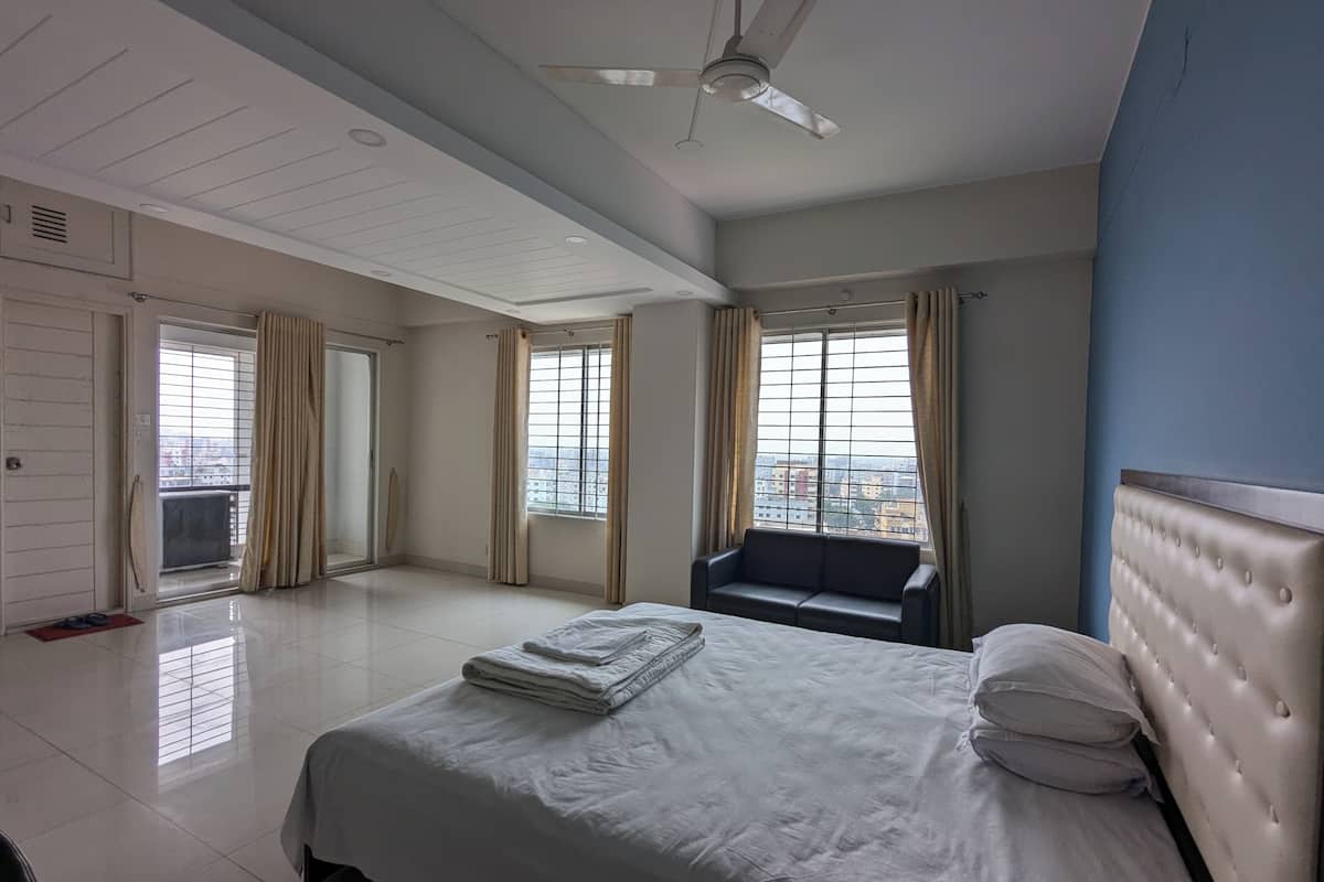 A hotel room with a bed, a chair, a ceiling fan, and large windows with curtains.