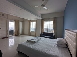 Quarto de hotel com cama, poltrona, ventilador de teto e janelas amplas com cortinas.