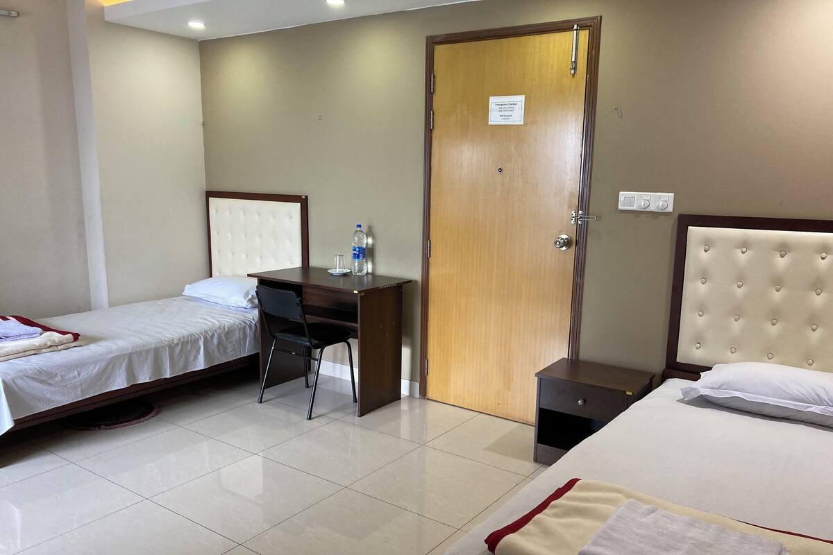 A hotel room with two single beds, a desk with a chair, a wooden door, and a small bedside table.