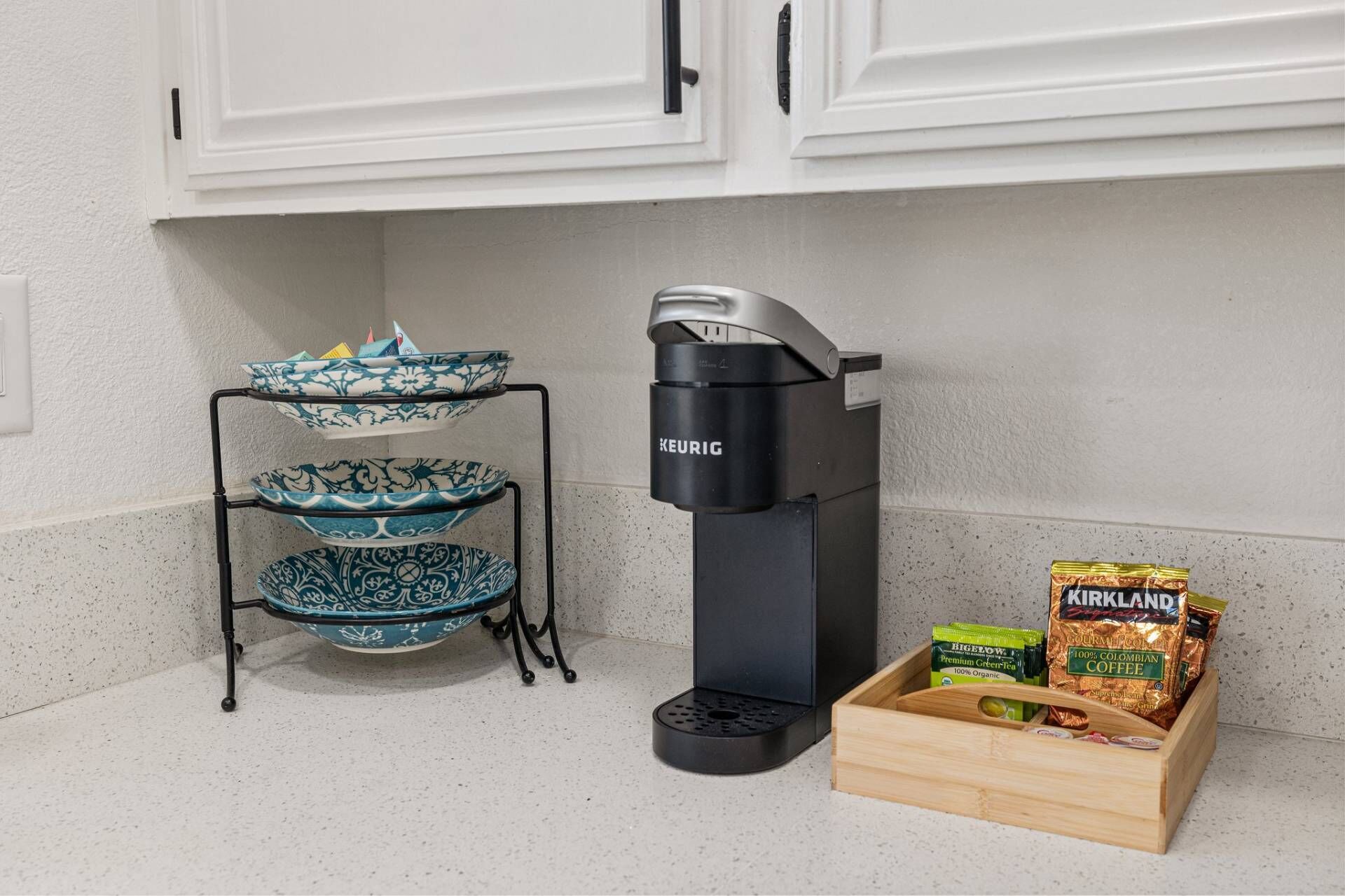 A kitchen area with a Keurig coffee maker, a three-tiered blue and white patterned bowl rack, and a wooden crate containing Kirkland coffee and green tea bags.
