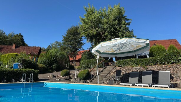 Een zwembad met een loungeruimte, een overdekte parasol en een trap die naar een gebouw met een rood dak leidt.