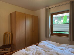 A bedroom with a wooden wardrobe, a single bed with white bedding, a chair, and a window with curtains.
