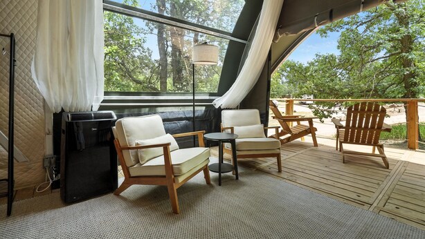 Un espace extérieur accueillant avec deux chaises et une petite table, un ventilateur de plafond et une vue sur les arbres à travers une grande fenêtre.